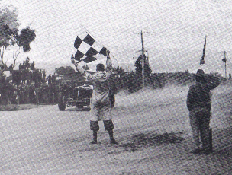 Black-and-white photograph of Peter Whitehead driving his ERA R10B racing car towards the chequered flag to win the 1938 Australian Grand Prix at Mount Panorama, Bathurst. A race official waves the flag in the foreground as Whitehead approaches, with spectators lining the dusty circuit behind.